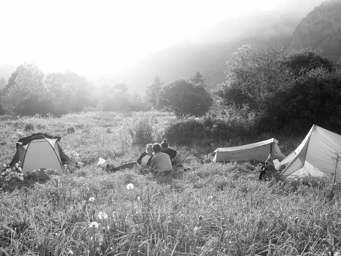 3 randonneurs assis dans l'herbe au milieu de 3 tentes installées dans une vallée en montage. Photo noir et blanc
